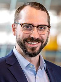 man with short hair and glasses smiling wearing blue collared shirt and jacket