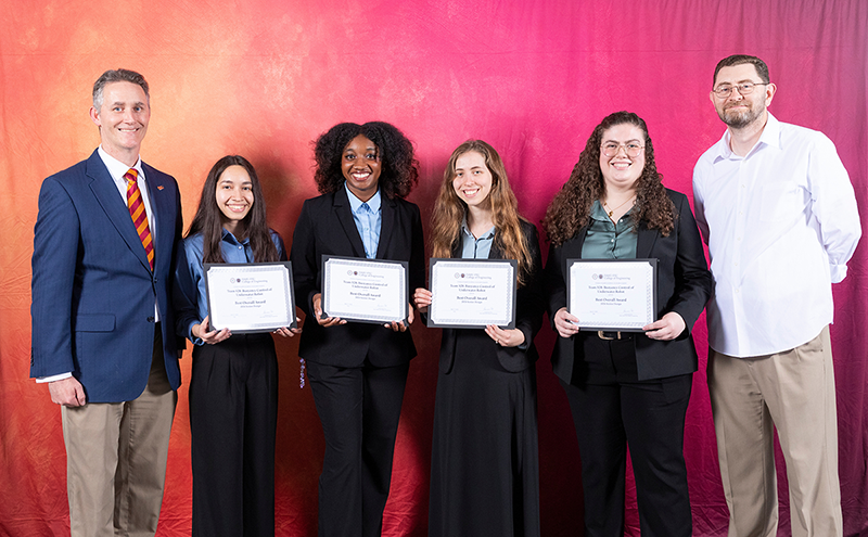 Four students in business attire stand in front of a red gradient backdrop holding certificates labeled “Best Overall Award,” flanked by two faculty members, all smiling toward the camera at an awards presentation.