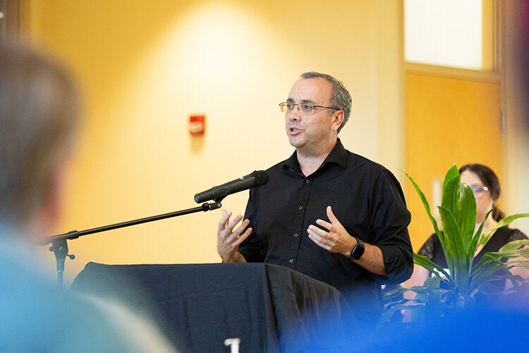 Man in a black shirt speaking at a podium with a microphone, gesturing with both hands in a well-lit indoor event space.
