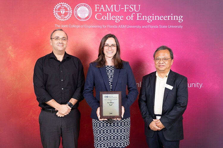 Three individuals pose in front of a FAMU-FSU College of Engineering backdrop, with the person in the center holding a “1st Place” award plaque.