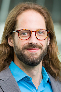 man with long hair and brown glasses in blue collared shirt and gray coat and beard smiling at camera