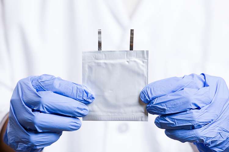 Close-up of a pair of latex-gloved hands holding a flat, square battery in front of a white lab coat.