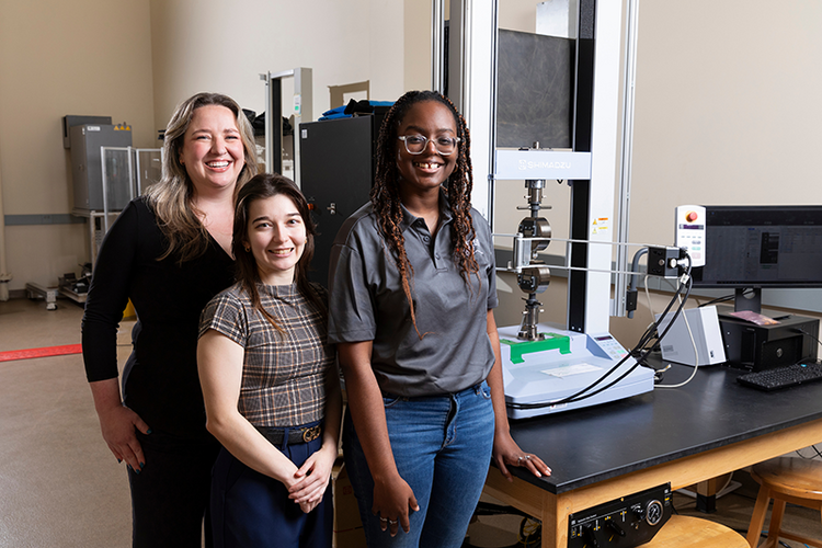 Three women posing in a lab next to a vertical materials testing machine mounted on a workbench, with stools, cables, and lab equipment arranged around the workspace.