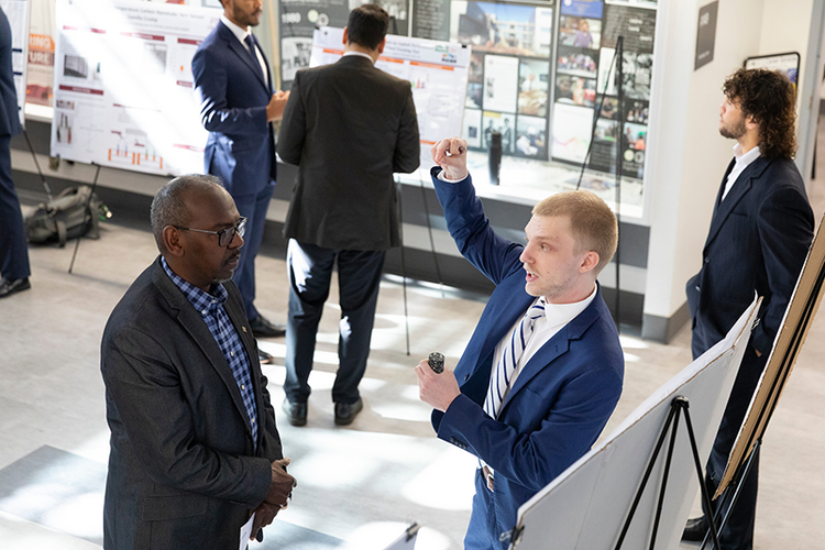 A man in a blue suit explaining a poster presentation to another attendee while pointing upward, with additional poster sessions in the background.