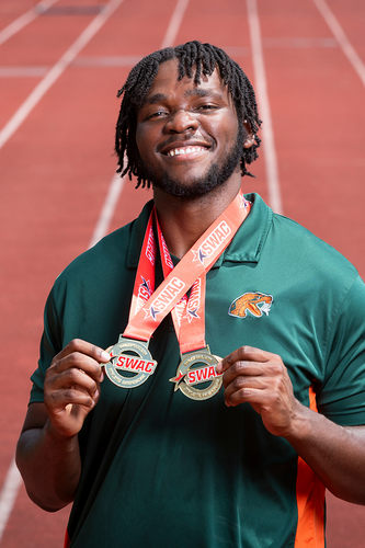 A student athlete smiles while holding two medals on a red outdoor track
