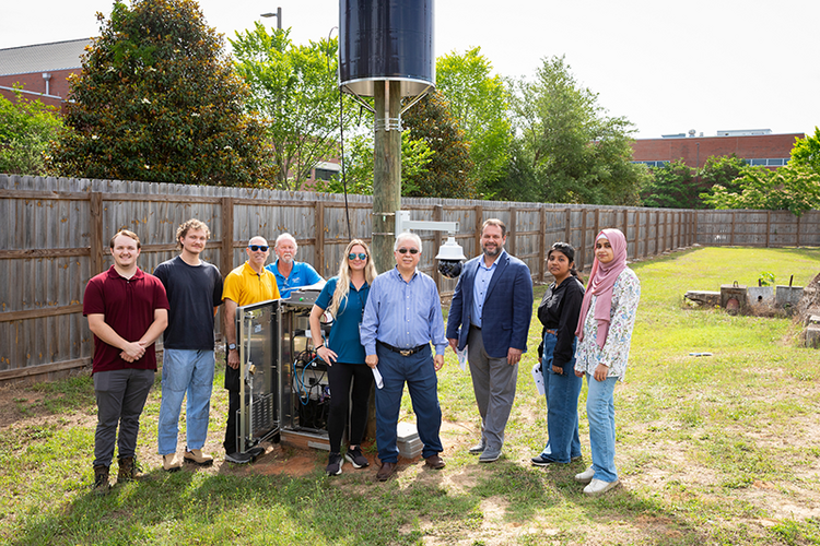Group poses outdoors beside a pole-mounted solar-wrapped device, open equipment cabinet, and mounted camera.