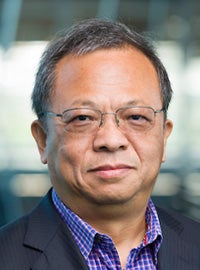 man with short gray hair and glasses wearing purple checked shirt and gray coat looks at camera