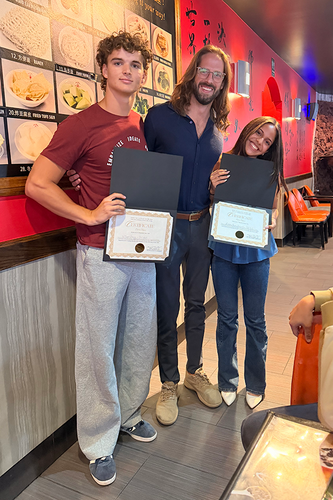 Two students hold certificates in black presentation folders as they pose with a smiling man in a restaurant. The student on the left wears a maroon T-shirt with “Empathize” and “Ideate” printed on it, and the student on the right wears a blue top and jeans. A menu board and red wall with Chinese lettering fill the background.