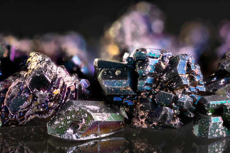Macro photo of an iridescent crystalline mineral specimen with angular, stepped crystal facets showing a rainbow metallic sheen in blues, greens, and purples against a dark, blurred background