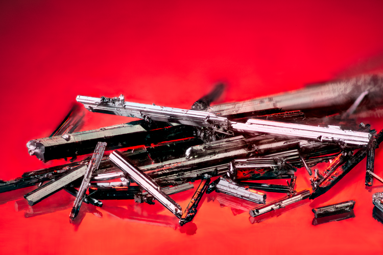 Macro photo of elongated, needle-like metallic crystal specimens with a bright silver mirror-like finish, scattered against a saturated red background