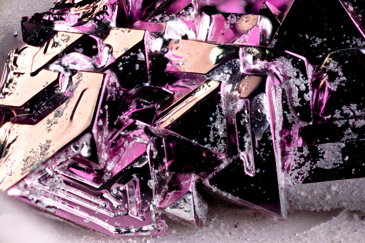 Macro photo of an AuAl2 (gold-aluminum) crystal specimen with angular, faceted crystal formations in deep purple and pink hues, dusted with fine white mineral particles.
