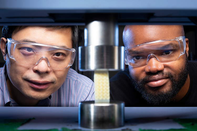 Two researchers wearing safety glasses observe a lattice foam sample positioned between cylindrical metal plates in a compression testing machine.