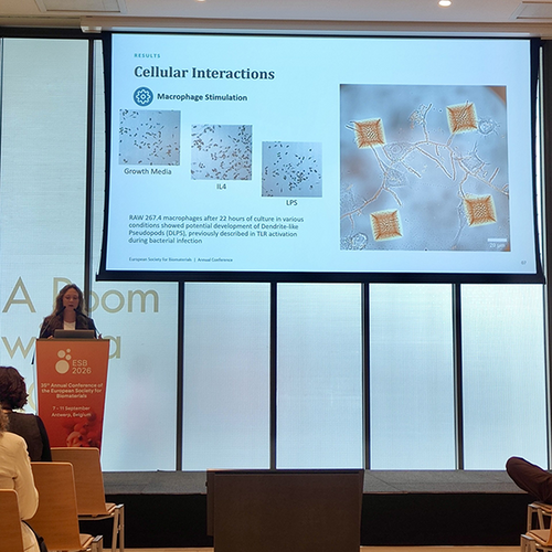 woman at podium talking at conference with medical presentation slide titled "cellular interactions" beside her