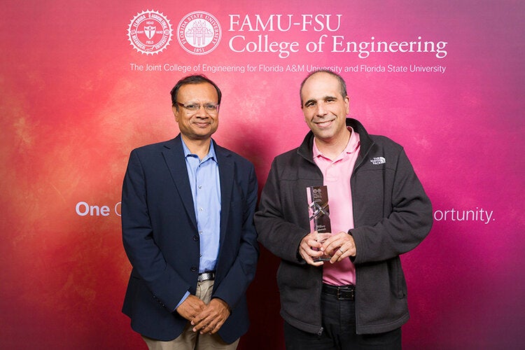 Two individuals stand in front of a FAMU-FSU College of Engineering backdrop, with the person on the right holding a clear glass award and smiling.