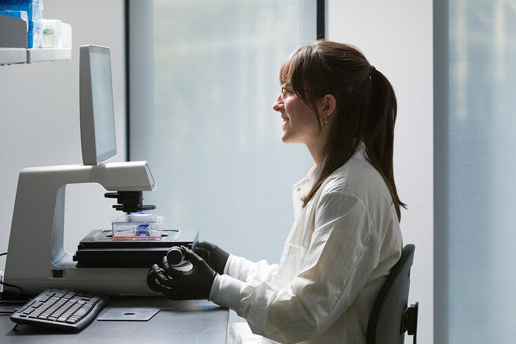 A woman in a lab works at a microscope with a screen.