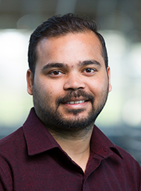 Headshot of a smiling man with short dark hair and a trimmed beard, wearing a maroon button-up shirt, positioned indoors with a softly blurred background