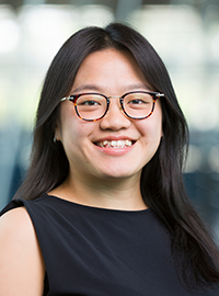 Portrait of a smiling woman with glasses and long dark hair wearing a sleeveless black top against a softly blurred indoor background.