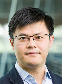 A professional headshot of a man wearing glasses, a navy blazer and a light blue button-down shirt, standing in front of a blurred indoor background.