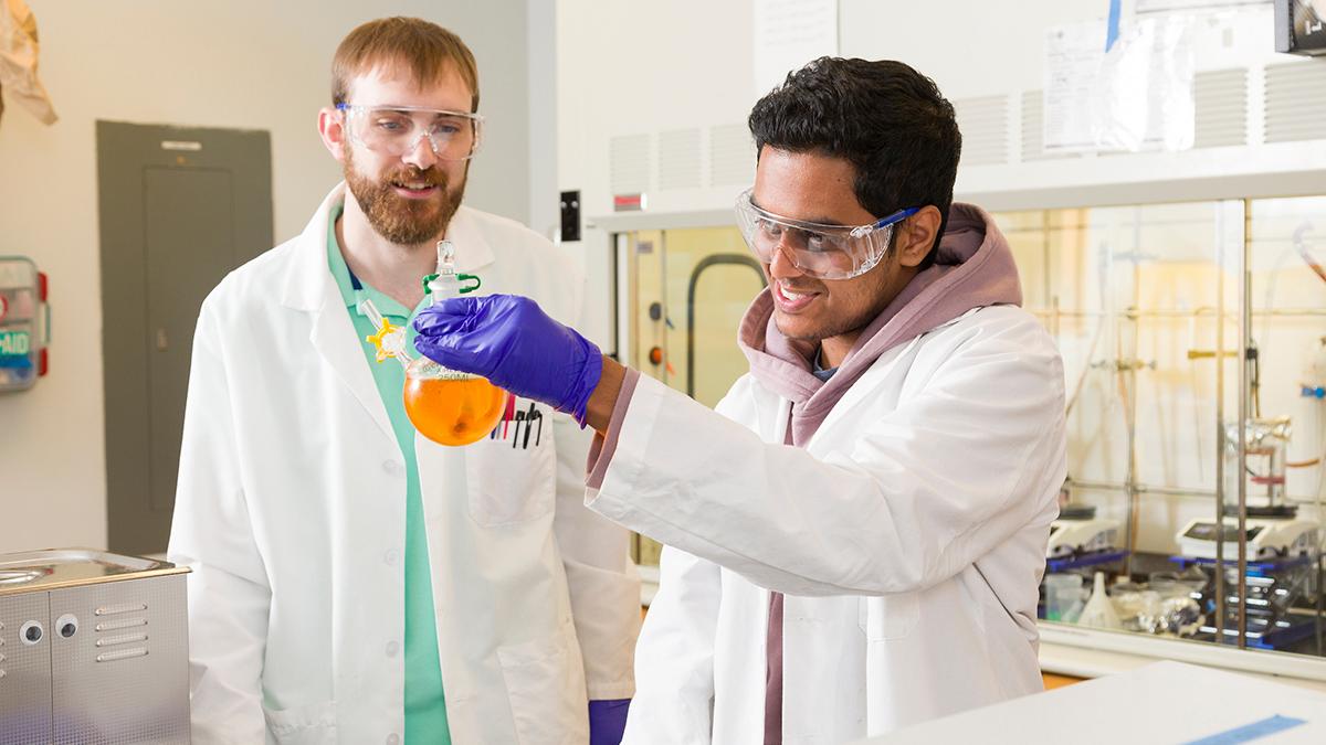 photo of two men in lab coats one holding beaker of orange fluid