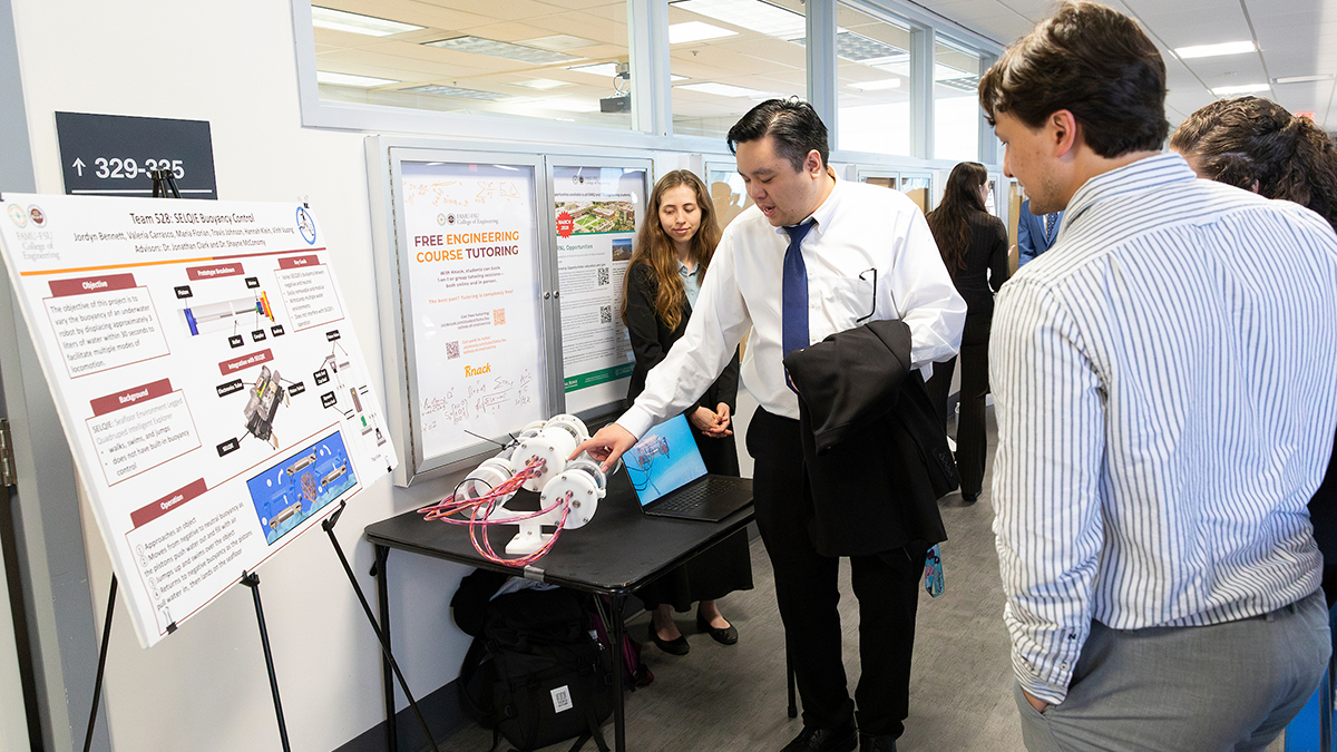 A student in a white shirt and tie demonstrates a cylindrical engineering device with attached wiring on a table while two attendees observe and ask questions, with a project poster on buoyancy control displayed beside them in a hallway setting.