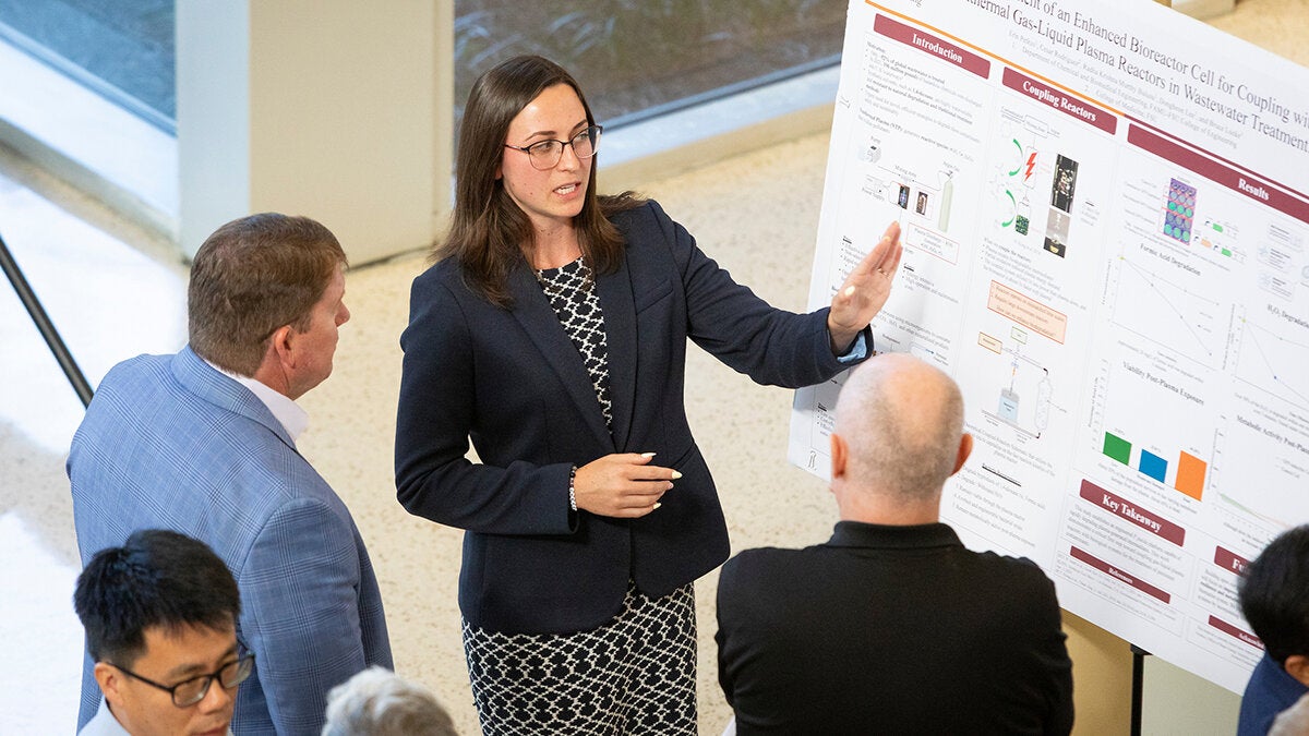 Presenter points to a section of a research poster while explaining findings to two individuals standing nearby.