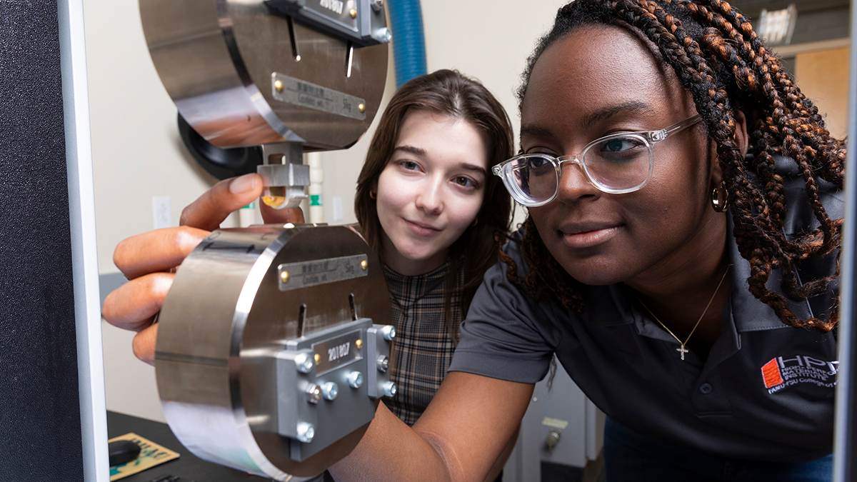 Two women closely observing and adjusting a materials testing device, with one reaching toward the sample mounted between metal grips while the other looks on.