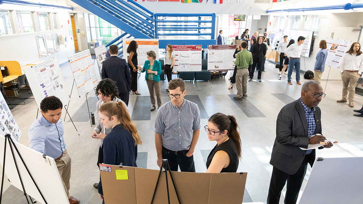 Overhead view of multiple groups of people discussing research posters arranged on easels in a bright atrium space.