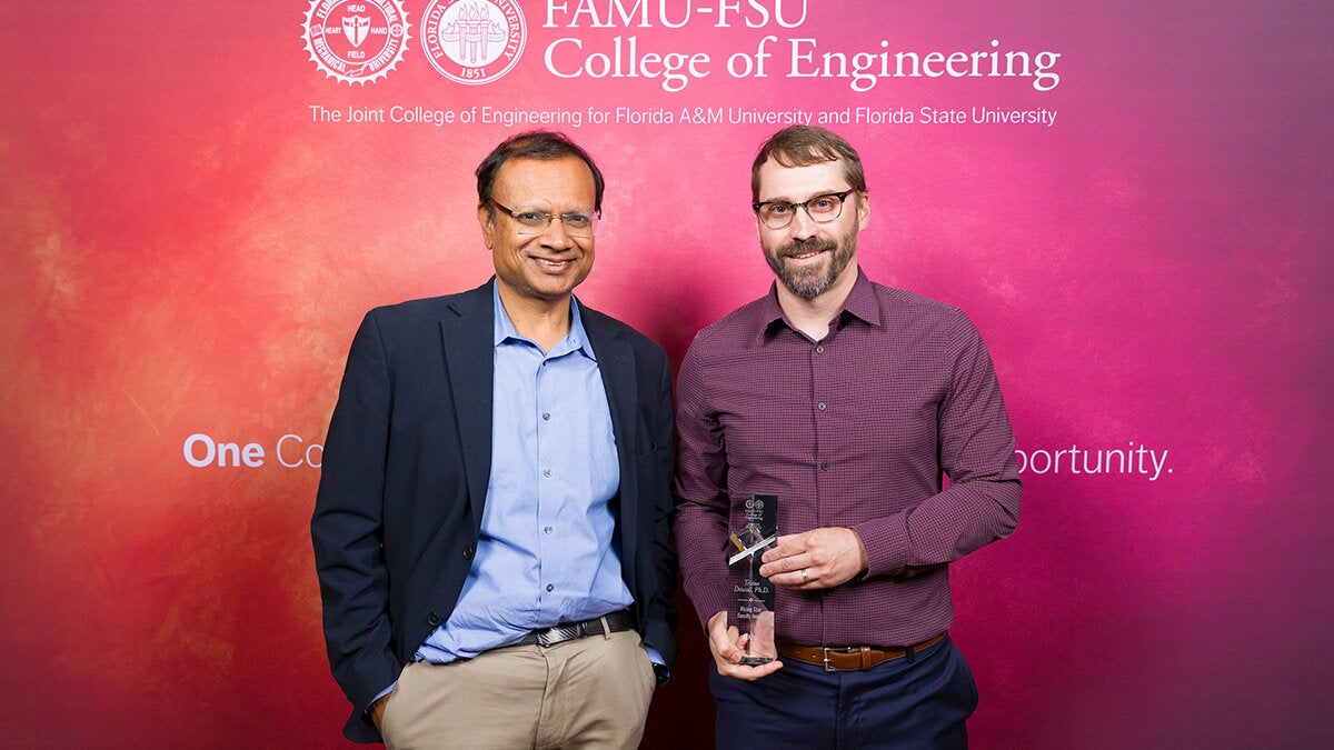 Two individuals pose in front of a FAMU-FSU College of Engineering backdrop, with the person on the right holding a clear glass award.