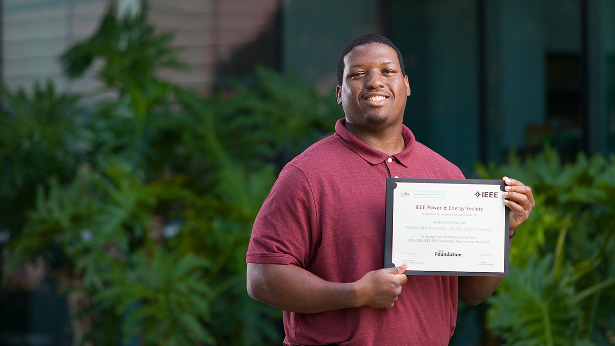 A student stands outdoors holding an IEEE Power & Energy Society scholarship certificate while smiling toward the camera.
