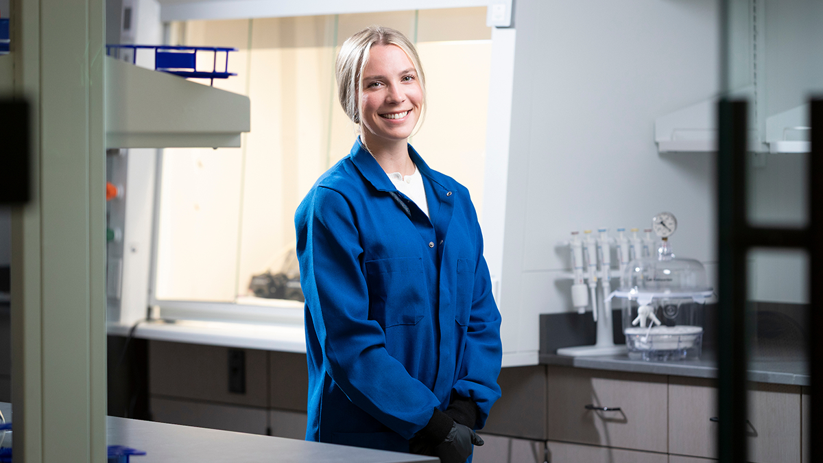 A researcher in a blue lab coat stands in a laboratory near a fume hood and lab equipment.