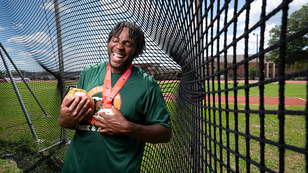 A student athlete laughs while holding a throwing weight and medals inside an outdoor throwing cage