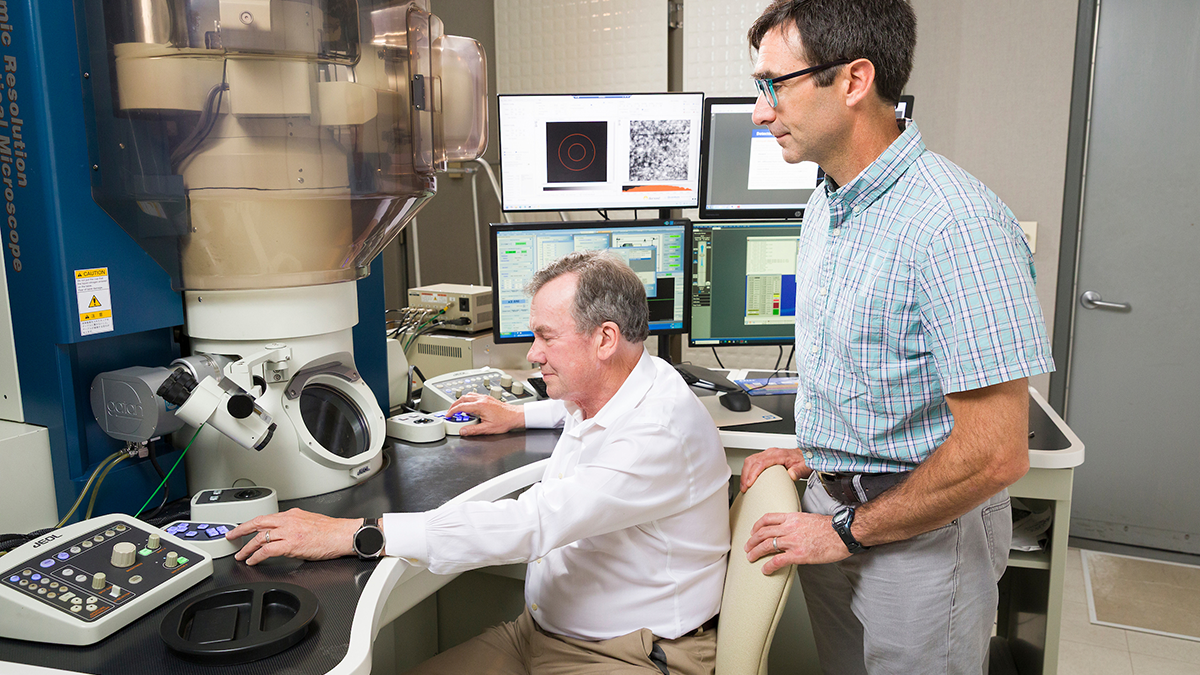 One researcher uses the controls of an atomic-resolution analytical microscope while another observes beside him.