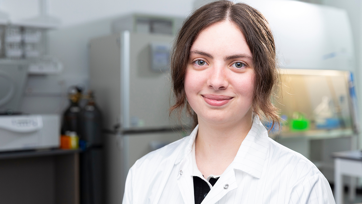 A student wearing a white laboratory coat sits in a research laboratory with scientific equipment visible in the background and smiles at the camera.