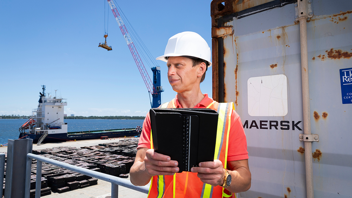 Man wearing a white hard hat and orange reflective safety vest holds an open black tablet while standing beside a shipping container at a waterfront cargo facility, with a ship and crane in the background.
