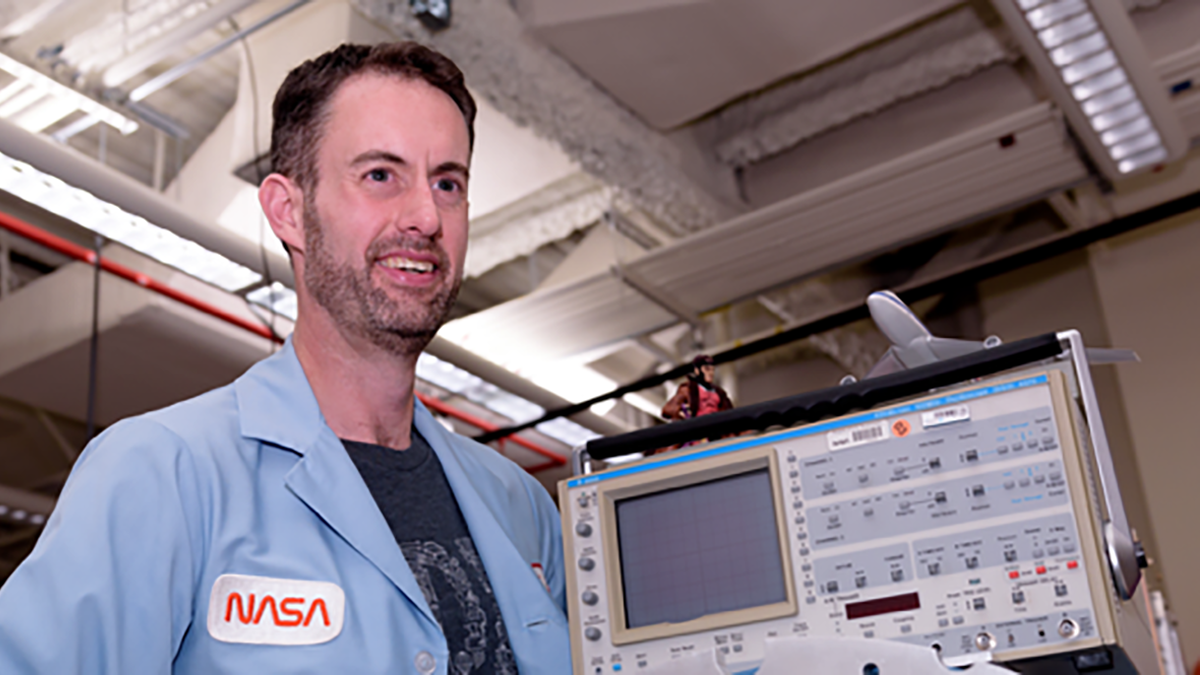 man in blue NASA shirt and beard smiling looking to right past camera
