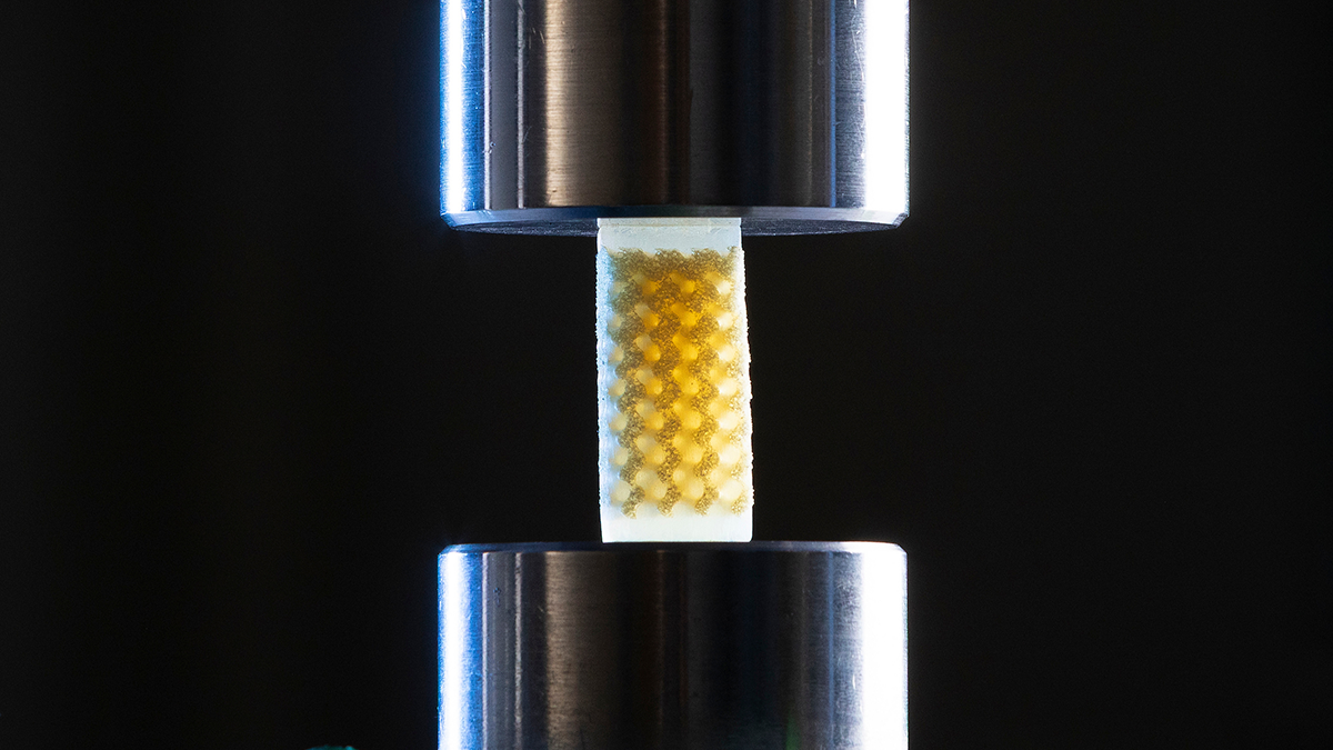 Close-up of a small lattice foam sample with a repeating internal structure positioned between two cylindrical metal plates of a compression testing machine.