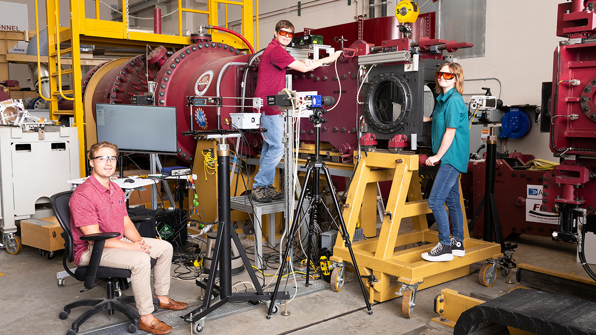Three students pose around a large red experimental apparatus in an engineering laboratory. One sits at left, one stands on a platform beside the equipment, and one stands on a yellow rolling platform at right.