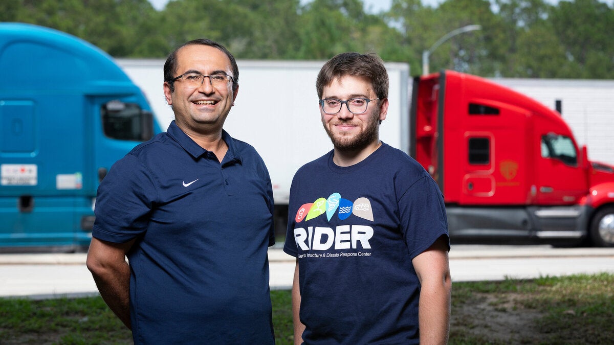 Two men stand outdoors in front of semi-trucks parked side by side, with trees and a clear sky in the background. The man on the right wears a navy RIDER T-shirt.