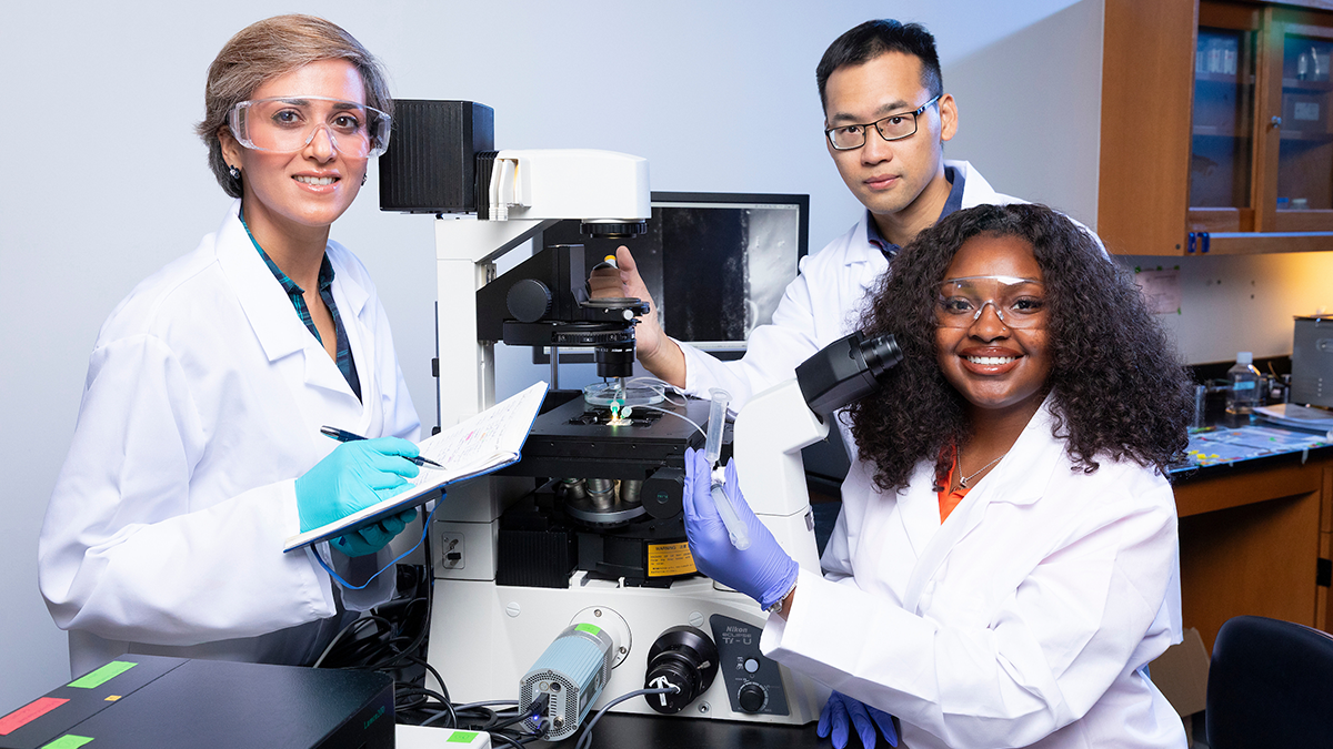 Three researchers wearing white lab coats and protective eyewear pose around a laboratory microscope. A woman on the left holds a notebook and pen, a man behind the microscope adjusts the equipment, and a woman on the right smiles while holding a clear sample tube.