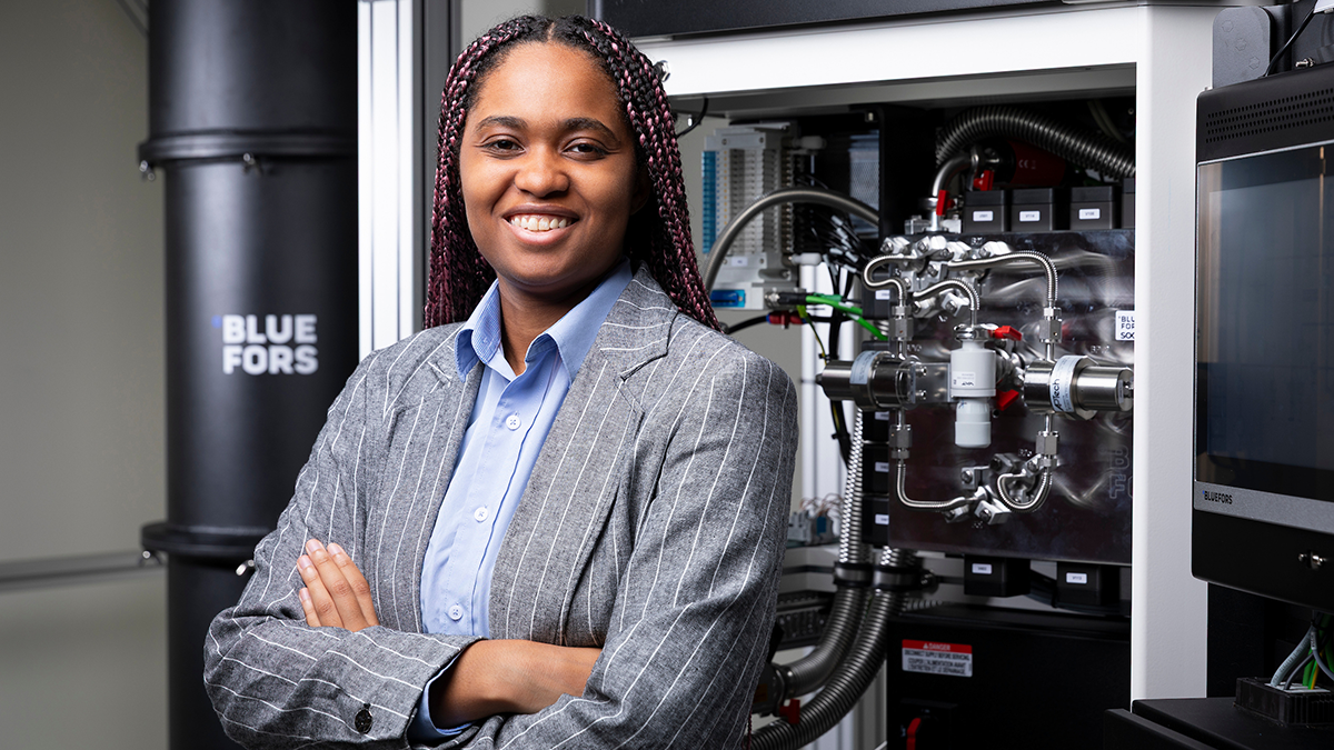 A smiling woman with long burgundy braids stands with her arms crossed in front of laboratory equipment, wearing a gray pinstripe blazer over a light blue shirt. Metal pipes, valves and a large black Bluefors cylindrical unit are visible behind her.