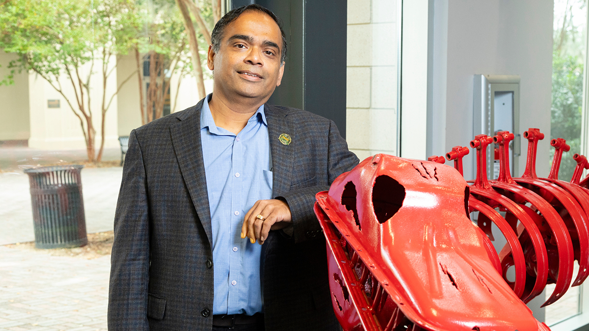 A man in a gray plaid blazer and light blue shirt stands smiling with one arm resting on a red metal sculpture of a dinosaur skeleton inside a building lobby, with a brick courtyard visible through the windows behind him.