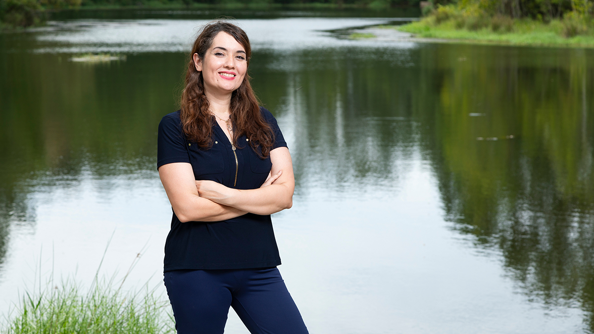 Woman in a navy blouse standing with arms crossed in front of a calm lake, smiling at the camera.