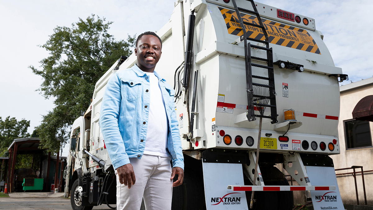 A smiling man wearing a light blue denim jacket and white pants stands in front of the rear of a large white garbage truck outdoors.
