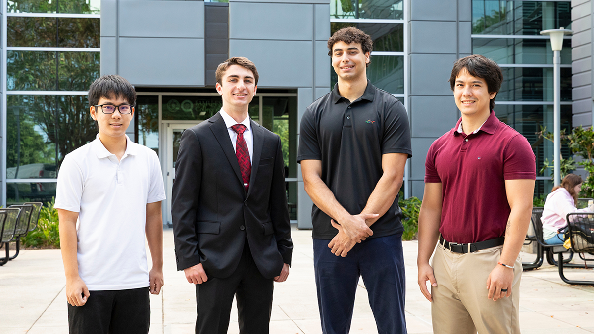 Four young men pose together in an outdoor courtyard in front of a modern glass-and-metal building.
