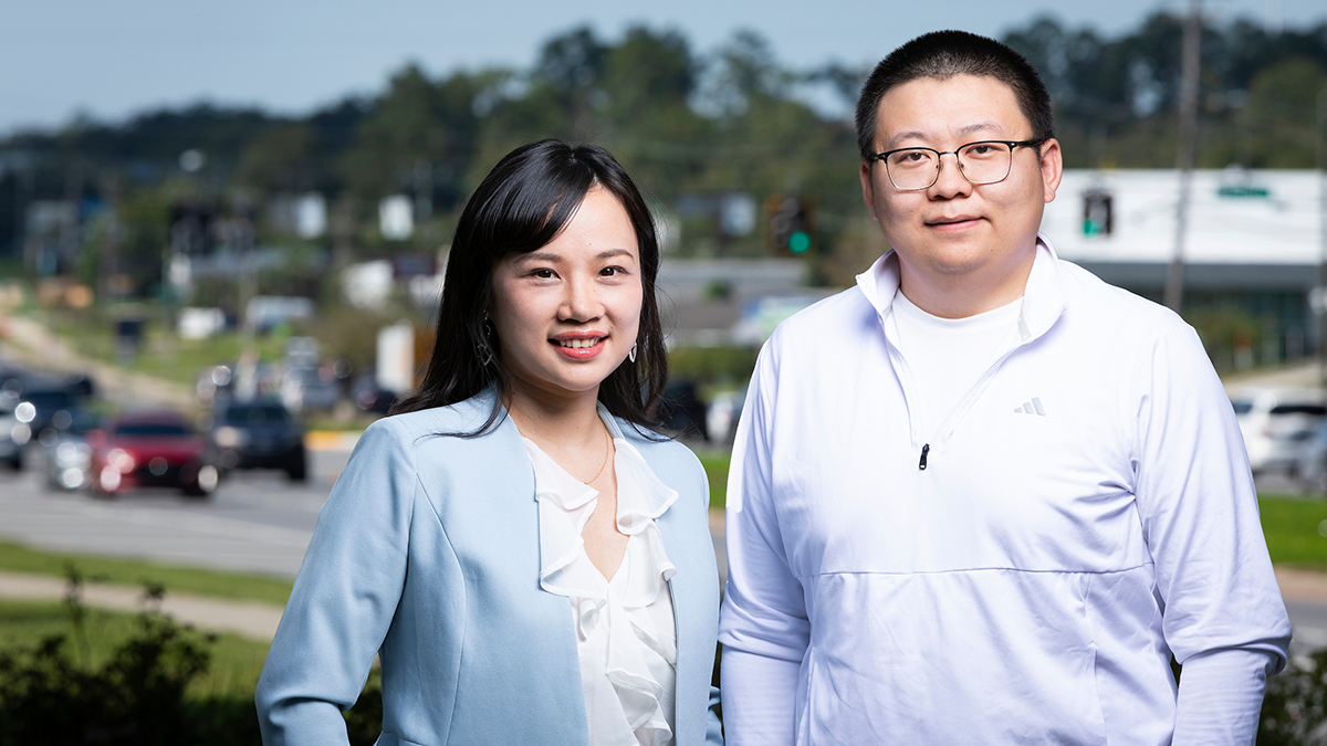 A woman with long black hair wearing a light blue blazer stands beside a man with short black hair, glasses, and a white quarter-zip pullover. Both smile at the camera, with passing vehicles and trees blurred in the background.