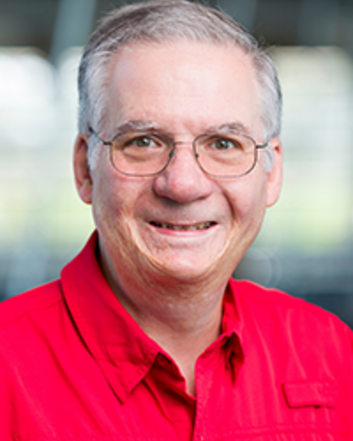 Portrait of a smiling man with glasses and gray hair wearing a red collared shirt against a softly blurred indoor background