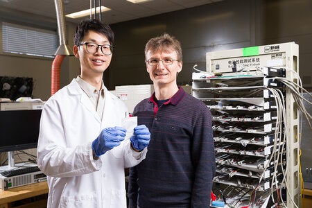 A researcher wearing a lab coat holds a flat, square battery while standing next to a professor in a laboratory.