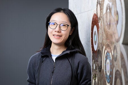 Portrait of a woman with long dark hair and glasses leaning beside a large circular patterned wall artwork, arms crossed with a neutral expression.