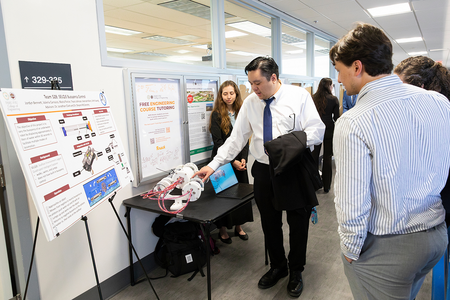 A student in a white shirt and tie demonstrates a cylindrical engineering device with attached wiring on a table while two attendees observe and ask questions, with a project poster on buoyancy control displayed beside them in a hallway setting.