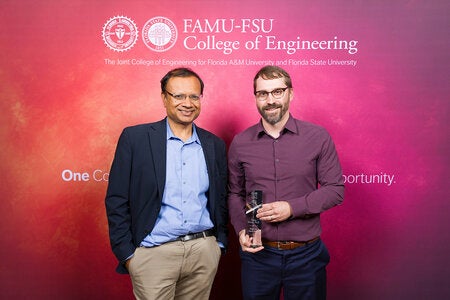 Two individuals pose in front of a FAMU-FSU College of Engineering backdrop, with the person on the right holding a clear glass award.
